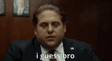 a man in a suit and tie is saying `` i guess bro '' while sitting at a table .