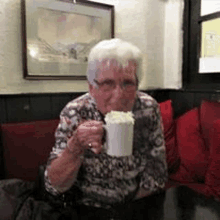 an older woman is sitting at a table drinking from a mug