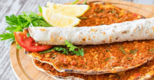 a stack of lahmacun pizzas on a wooden cutting board with lemon slices and tomatoes .