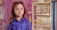 a little girl in a blue shirt is standing in front of a shelf with a lot of glassware on it .