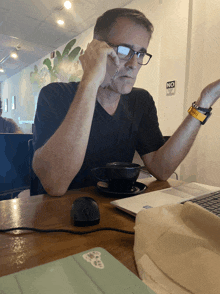 a man wearing glasses sits at a table with a cup of coffee and a laptop