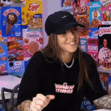 a woman wearing a hat and a black shirt is sitting in front of a wall with cereal boxes on it .