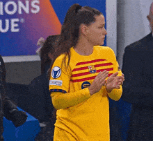 a female soccer player wearing a yellow jersey is clapping her hands in front of a sign that says champions league