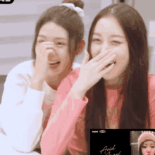 two girls are laughing and covering their mouths with their hands while sitting next to each other .