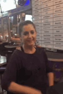 a woman in a black shirt is smiling in front of a wall with a list of beers on it