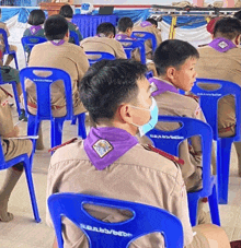 a man wearing a mask sits in a row of blue chairs with a purple scarf on his neck