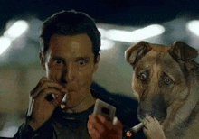 a man is smoking a cigarette while holding a cell phone and a dog is standing next to him .