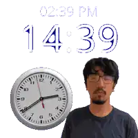 a man wearing glasses stands next to a clock that says 14:39