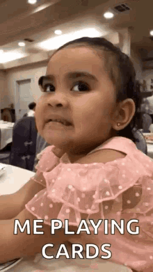 a little girl in a pink dress is sitting at a table playing cards and making a funny face .