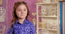 a young girl in a blue shirt is standing in front of a shelf filled with trophies and crowns .