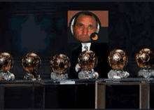 a man in a suit is standing in front of a row of soccer trophies .