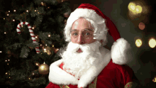 a man dressed as santa claus stands in front of a christmas tree with a candy cane hanging from it