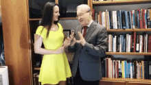 a man in a suit and tie is talking to a woman in a yellow dress in front of a bookshelf