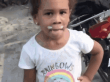 a little girl wearing a rainbow shirt is standing in front of a motorcycle and making a funny face .