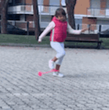 a little girl in a pink vest and white pants is playing with a pink ball on a brick sidewalk .