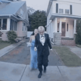 a woman in a blue dress and a man in a tuxedo walk down a sidewalk