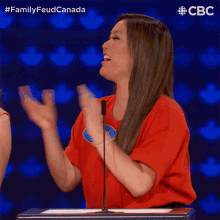 a woman in a red shirt stands in front of a microphone with her arms in the air and the words family feud canada behind her