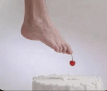 a person 's foot is reaching for a cherry on top of a cake .