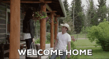 a man in a cowboy hat stands on a porch with a woman holding a bag and the words welcome home above him