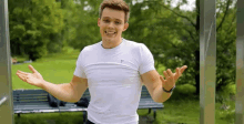 a young man in a white shirt is standing in front of a park bench with his arms outstretched .