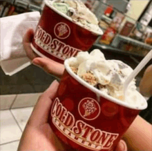 a person is holding two cups of cold stone creamery ice cream with spoons .