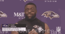 a man with a beard is standing in front of a purple wall with a ravens logo on it