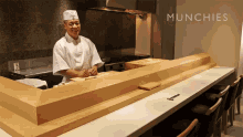 a man in a chef 's uniform stands behind a counter with the words munchies written on the wall behind him