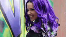 a woman with purple hair is smiling in front of a wall with the letter v on it