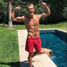 a shirtless man in red shorts flexes his muscles by a pool