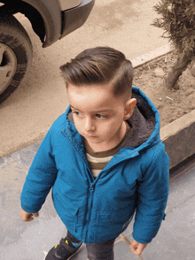 a young boy wearing a blue jacket with a hood stands in front of a car