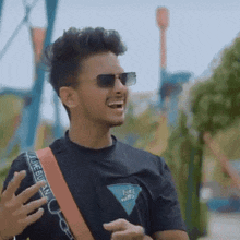 a young man wearing sunglasses and a black t-shirt is laughing while standing in front of a roller coaster .