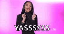 a woman is standing in front of a pink background and making a heart shape with her hands with the words yassss written on it .