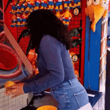 a woman in a blue shirt is playing a game in front of a wall of stuffed animals including one that says simpson