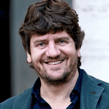 a man with a beard is smiling for the camera while wearing a blue jacket and a blue shirt .