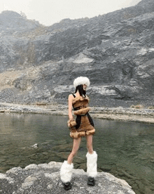 a woman in a furry dress and furry boots is standing on a rock near a lake .