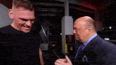a man in a suit talks to another man in a black shirt in a dark room