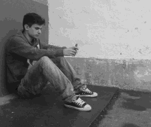 a black and white photo of a young man sitting against a wall looking at his cell phone