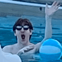 a man wearing sunglasses is swimming in a pool while holding a blue beach ball .