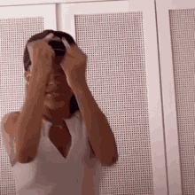 a woman in a white tank top is putting on a hat in front of a white wall .