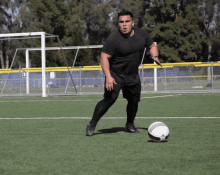 a man in a black shirt is kicking a soccer ball on a field with the number 3 on the fence behind him