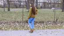 a woman wearing a green sweater and blue jeans is walking down a sidewalk next to a chain link fence .