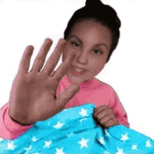 a woman in a pink shirt is waving her hand while laying in a bed with a blue blanket with white stars on it .