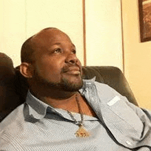 a man with a beard is sitting in a chair wearing a blue shirt and a gold necklace .