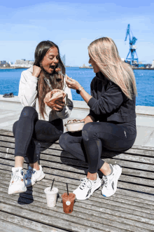 two women are sitting on a wooden bench eating food