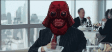 a man in a suit and tie with a red mask on his face is sitting at a table with a martini .