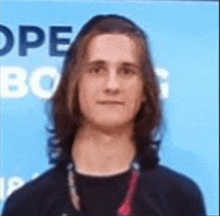 a young man with long hair is standing in front of a blue background and looking at the camera .