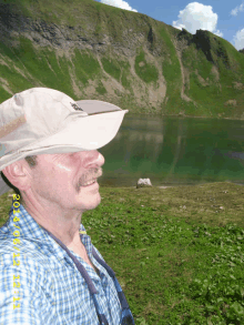 a man wearing a hat stands in front of a mountain and a lake