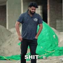 a man with a beard is standing in front of a pile of dirt and a green tarp with the word shit written on it