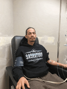 a man wearing a saskatoon sweatshirt sits in a chair