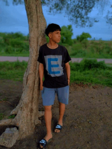 a young man leaning against a tree wearing a shirt that says e